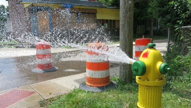 Yellow and green fire hydrant spraying water on a hot June day. Orange street cones block the corner sidewalk allowing for people to access the water. 