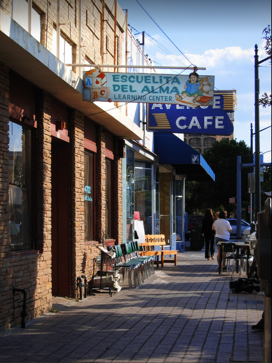 A street scene on Congress Avenue. A brown brick building has a blue sign hanging over the sidewalk saying, "Escuelita Del Alma Learning Center." The sign has a painting of a young child reading a book next to a teddy bear on one side. Playing blocks with the letters A, B and C are on the other. Chairs line the sidewalk outside the day care. Behind the Escuelita sign is another retro 50s sign partially obscured that says, "Avenue Cafe." A couple people are milling about down the block.