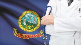 A person in a medical coat holding a stethescope, seen from shoulders to hips. The background is of the Idaho state flag.