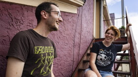 Two people who look to be in their 30s laugh while standing and sitting on the stairs outside a burgundy building.