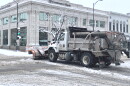 The side and back of a City of Columbia snowplow is visible as it pushes snow with its orange blade through an intersection in downtown Columbia.