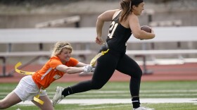 Woman running with a football on a field as another woman lunges to pull a yellow flag attached to the other woman's waist. 