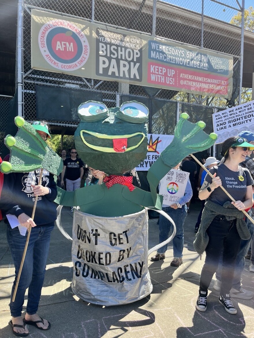 A group of people dressed up in a frog costume with raised arms stand in a crowd at a protest. The frog’s body reads, “Don’t get cooked by complacency.”