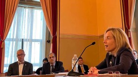 Caitlin Halligan sitting at a desk. In the background, people look on as she answers questions.