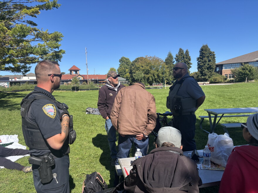 Traverse City Police Officer Matthew Kirkey (left) and Police Support Services Worker Brad Neuder (right) explaining the department's new enforcement efforts to a table of homeless individuals at Hull Park (Credit: IPR News / Maxwell Howard)