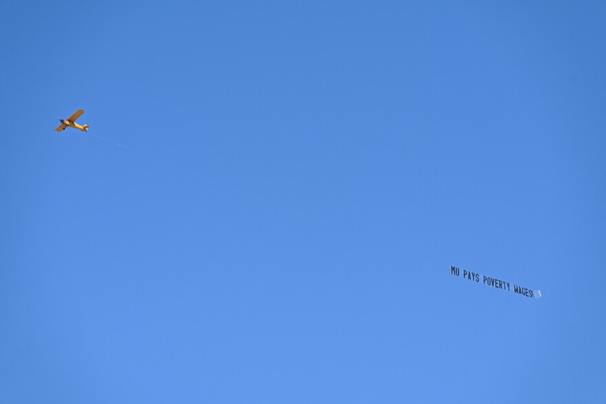 A plane flies with a sign saying “MU PAYS POVERTY WAGES” on Thursday on the University of Missouri campus.