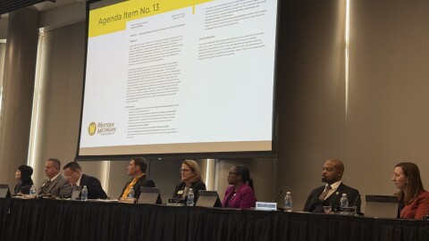 The Western Michigan University Board of Trustees sits at a long, black table, each with a microphone in front of them. The trustees are all looking off to the side, and a projector behind them shows a document titled "Agenda Item No. 13". The agenda item is for the transfer of the Asylum Lake Preserve to Kalamazoo County. 