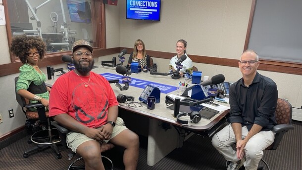 Five smiling people sit around a table in a radio talk studio: a woman front left has short curly brown hair and is wearing glasses, gold jewelry, and a green dress with a black belt; a man front center has a black mustache and beard and is wearing glasses, a brown baseball hat, a red spiderman t-shirt, a gold chain and pendant, khaki shorts, white socks, and black sneakers; a woman back left has long brown hair and is wearing sunglasses and a black and white dress; a woman back center has light hair in a braid and is wearing a blue dress with a design; a man at right has very short dark hair and is wearing glasses, a black button-down shirt, grey linen pants, and grey and blue sneakers