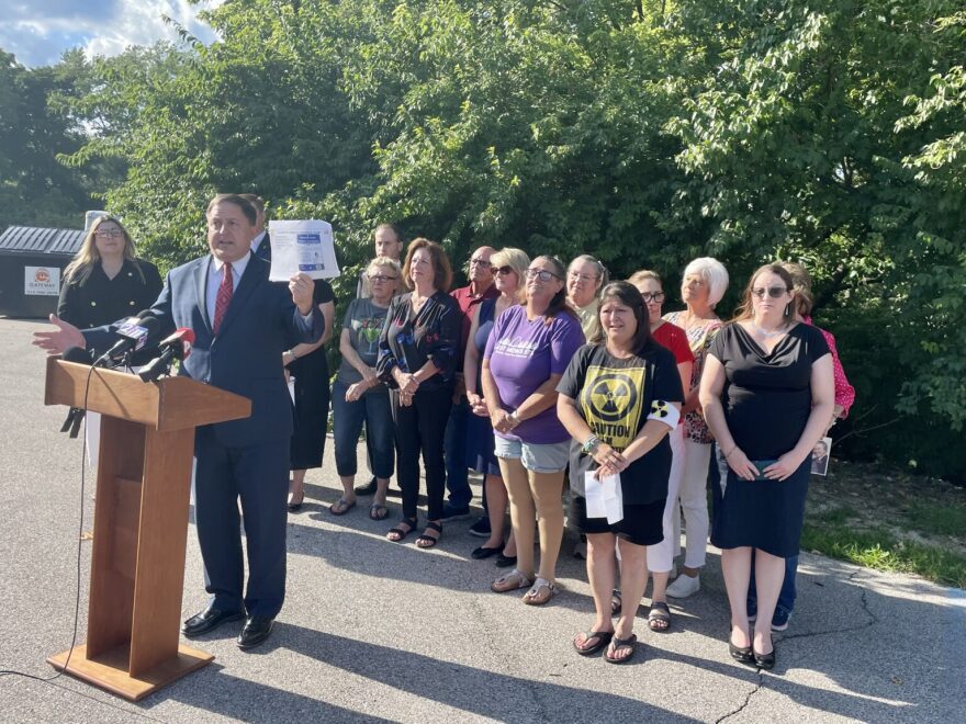 Secretary of State Jay Ashcroft stands at a podium on asphalt outdoors. A small crowd of people stands behind him, including one person with a radioactive symbol on their shirt.