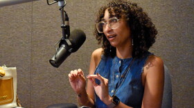 Woman in glasses and a blue denim top gestures while speaking into a microphone in a professional radio studio.