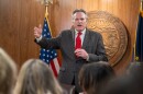 Man in wood-paneled room wearing a suit standing in front of state seal and flags