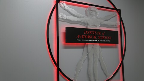 A sign showing Leonardo da Vinci's The Vitruvian Man, with text that reads 'Institute of Anatomical Sciences - Texas Tech University Health Sciences Center.' It has red lights glowing from behind it and onto the white wall.