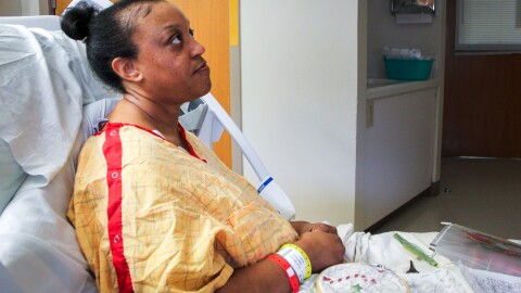 Shanice Jordan sits in a hospital bed in a patient room. She wears a yellow hospital gown with a light grid pattern on it and three hospital bracelets -- one red, one white and one yellow. She had a blanket over her lap, and on top of the blanket is some white fabric in an embroidery hoop with some progress made on stitching. The design includes several iconic Christmas symbols, like candy canes and holly. Jordan is a Black woman with her hair pulled into a bun.