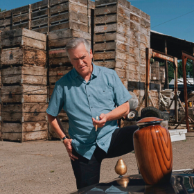 Bruce Campbell in a still from Ernie & Emma, smoking a cigarette while looking down at an urn placed on the hood of a car. There's a golden pear as the hood ornament.