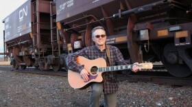 A man with a guitar kneels in the gravel alongside freight train cars