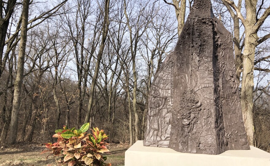 A large bronze sculpture featuring symbolism of indigenous culture and heritage stands alongside a colorful plant with trees in the background.   