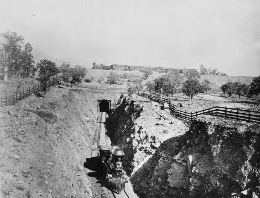 Photograph of two trains below and above on the Tehachapi loop, ca.1890. At center, a locomotive engine can be seen coming trhough a tunnel in the side of a hill, making its way through a shallow canyon. In the distance, a second engine can be seen pulling five cars in its train, moving across the ridge of the hill. Post-and-rail fence can be seen lining either side of the canyon.