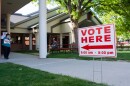 A "vote here" sign staked in the grass in front of some red picnic tables.