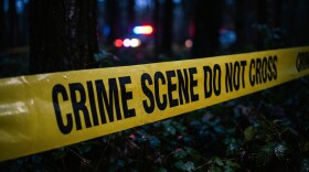 Yellow crime scene tape with the message "Crime Scene Do Not Cross" blocks a wooded area with police cruiser lights in the background