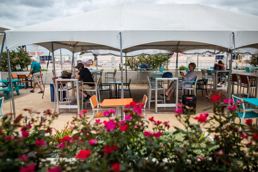 People dining outside under a white tent at the South Terminal. Blue and turquoise tables and chairs are set up. In the foreground are pink flowers.