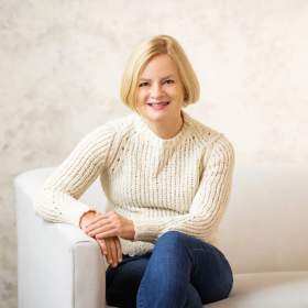 A woman with short blonde hair wears a white sweater and blue jeans and sits on a white couch. She crosses her hands and smiles at the camera. 