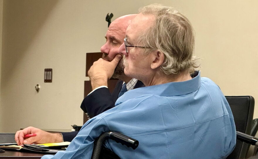 A man in a blue shirt is sitting in a courtroom in a wheelchair. His attorney is sitting to his right.