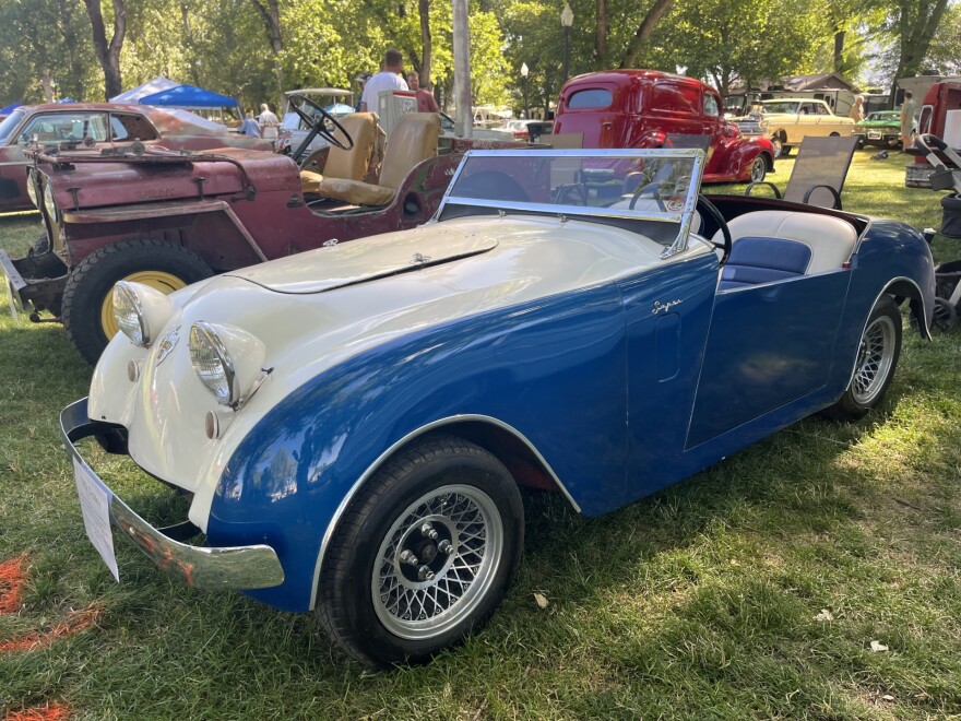 An old blue and white convertible sports car from the 1950s sits on grass with other old cars near it and in the distance to the left.