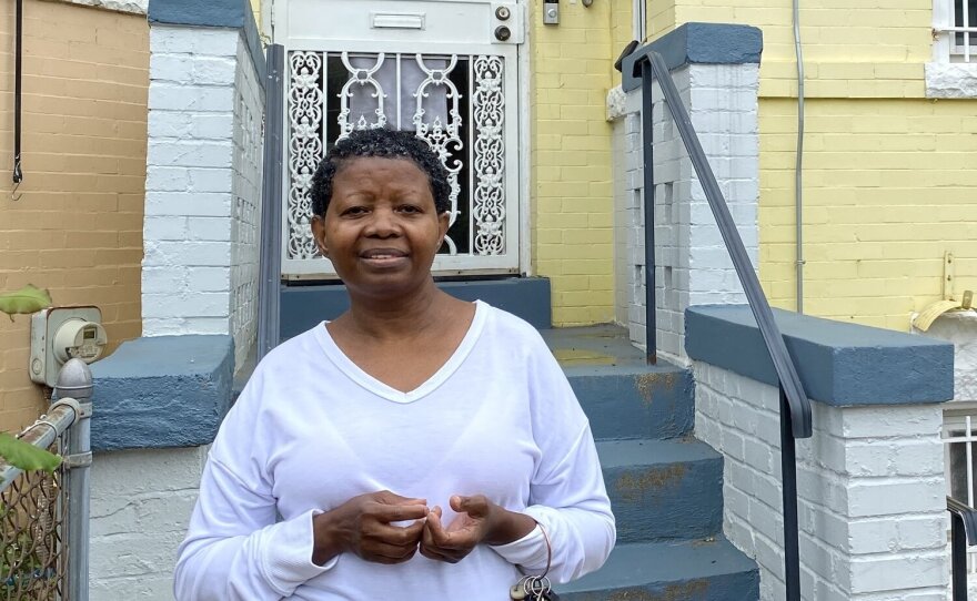 A woman wearing a white long-sleeved shirt stands half-smiling in front of a yellow house.
