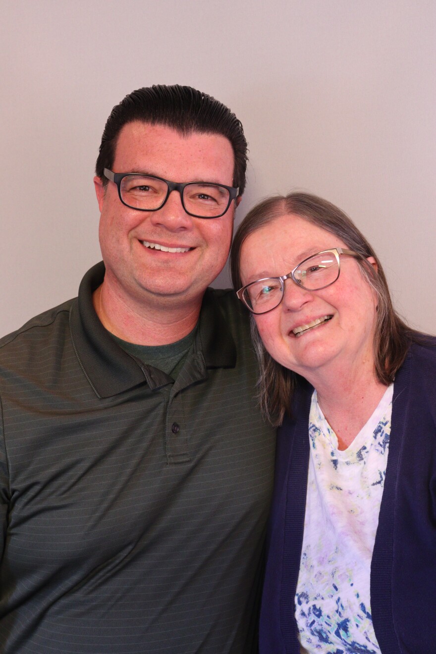 David and Cindy  Cordero side hug at their StoryCorps interview. David stands at least half a head taller than his mother, who leans her head in toward his shoulder. David has short dark hair and wears dark framed glasses and a dark heathered gray pullover.  The pullover is slightly unzipped and he is wearing a black tee shirt underneath it.  Cindy has light brown strawberry hued hair that falls to her shoulders.  Wisps of gray fall in front of her ears.  She has cat's-eye glasses.  She wears a blue printed white tee shirt and a dark cardigan.