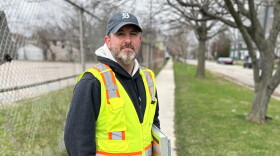 Man in a ballcap and greenish-yellow safety vest with clipboard under one arm smiles subtly while facing the camera