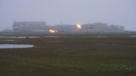 Flames seen above and in front of a large industrial facility on a foggy plain
