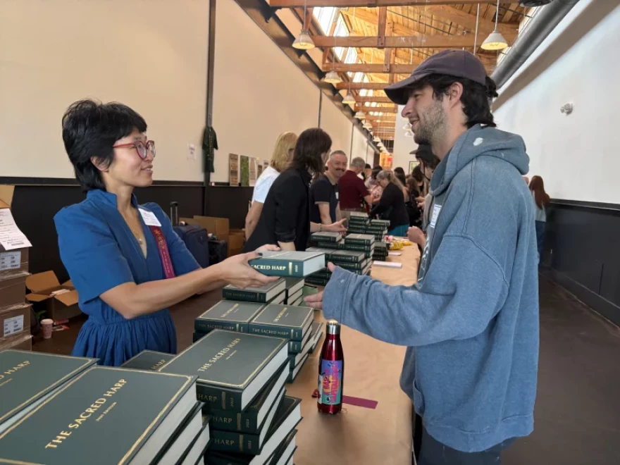 Leigh Cooper of San Francisco hands a brand new 2025 edition of The Sacred Harp to a singer at the book sales table just inside the convention entrance.