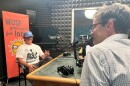 Man in white graphic t-shirt and blue hat with a moustache sits in chair in front of pink sign saying WUSF Your Spot for Local. A microphone is in front of him. The back of another man speaking into a microphone is in front of him. They're both siting at a table talking. 