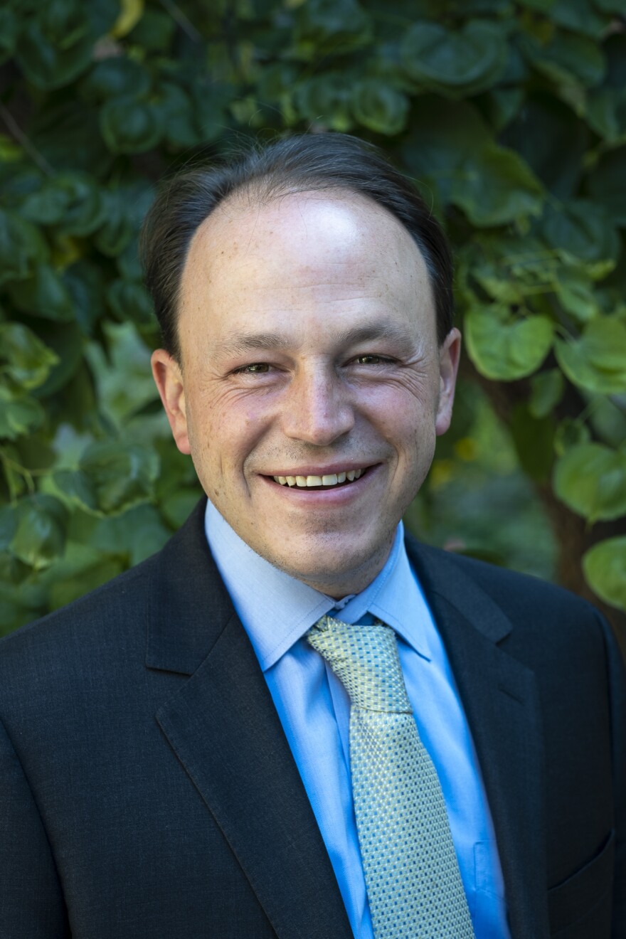 A middle aged white man smiles at the camera for a headshot. He's wearing a dark blue suit jacket with a light blue shirt and yellow and blue tie.