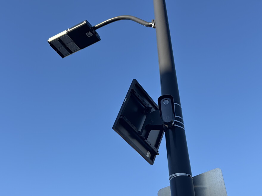 A security camera is affixed to a light pole. The photo is a worm's eye view. The camera has a very small solar panel above it, also affixed to the pole. 