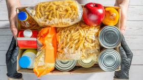Dos pares de manos, uno con guantes, sostienen una caja de cartón llena de pasta, fruta, conservas y leche.