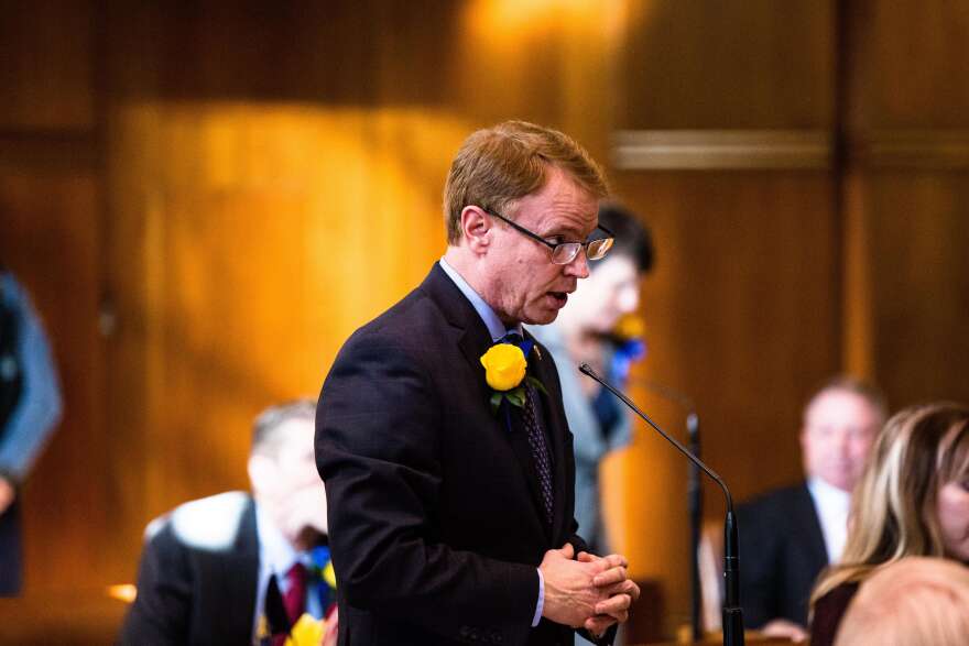 Tim Knopp is speaking at a microphone on the Senate floor. He is wearing a flower on his suit. 