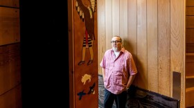 A man stands against a wooden wall and looks at the camera. One panel has a painting of a Native person in regalia. 