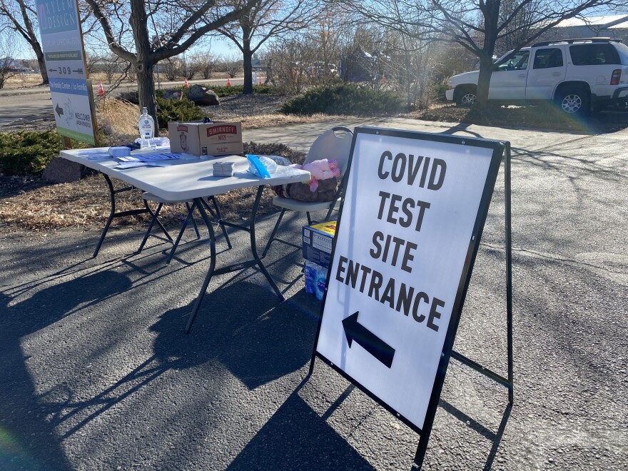 A sign in a parking lot next to a table reads, "COVID Test Site Entrance" with an arrow pointing to the left.