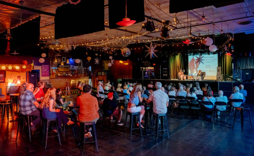 A large audience sits inside a music venue watching two speakers onstage beneath string lights and hanging decorations.