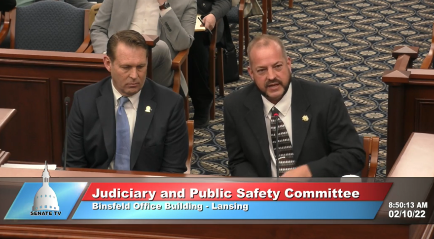 Randy Gilbert (left) is sitting on a wooden table inside a meeting room at Michigan's Capitol building. Gilbert is sharing his testimony to the Senate Judiciary Committee. Sitting next to him (left) is Eaton County Prosecuting Attorney Douglas Lloyd. Both are wearing a suit and tie. 