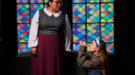 A young woman in a light blue headband kneels with praying hands in front of a woman who looks down at her. They are posed in front of a stained glass window.