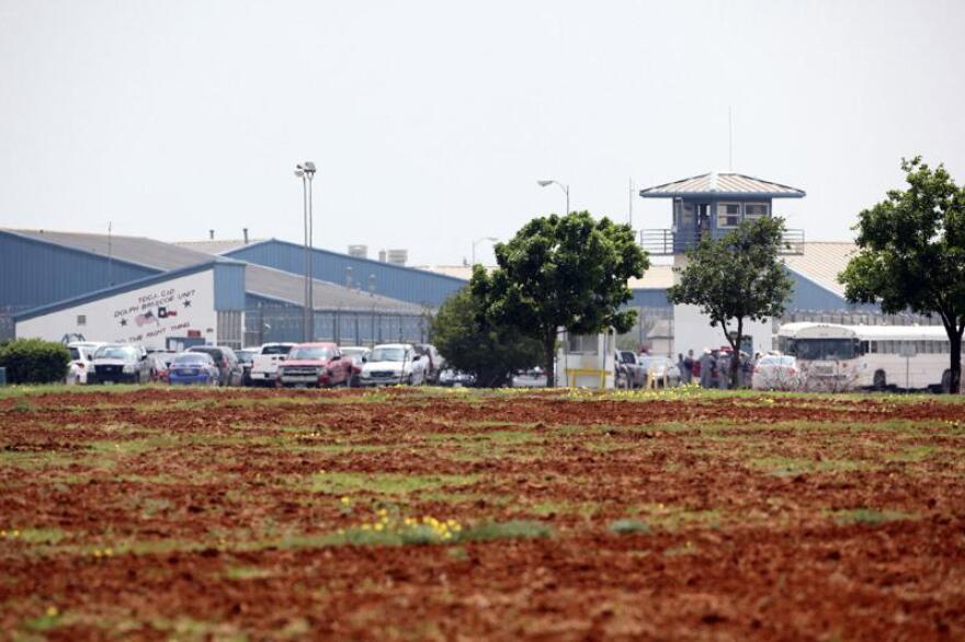 A gray metal group of buildings stands surrounded by fencing topped with razor wire. A guard tower stands near the right of the group of buildings.