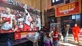 People mill about the interior of a large building. There is a giant TV monitor with Kansas City Chiefs football players displayed and a banner that reads "Chiefs Kingdom."