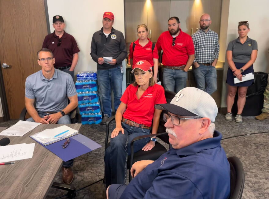 Three government officials sit at a desk, one man on the left, a woman in red in the middle, and a man wearing a baseball cap on the right.