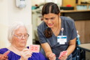 A nurse in scrubs leans over while holding playing cards next to a nursing home resident, also holding playing cards.