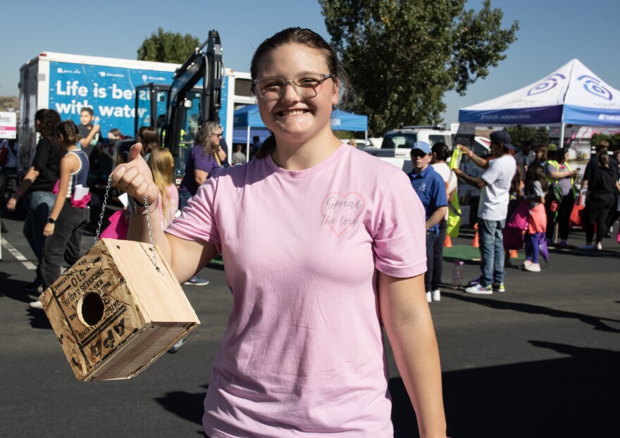 A girl in a pink t-shirt holds up a wooden handmade birdhouse and smiles.