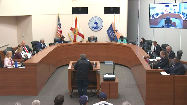 large meeting room with long dais with people sitting at it and a podium in front with a person speaking 