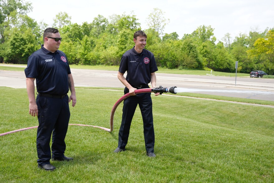 a man holds a fire hose while a more experienced firefighter watches