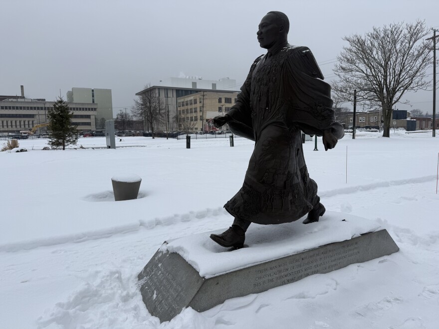 A statue of Martin Luther King Jr. is shown on a snowy day, and construction can be see in the background of the photo. In the statue, Dr. King is walking forward while wearing clerical robes that flow around him as he walks. On the robes, the faces of other civil rights leaders are etched alongside scenes like civil rights marches. A quote can be seen on the base of the statue that reads: “EVERY MAN MUST DECIDE WHETHER HE WILL WALK IN THE LIGHT OF CREATIVE ALTRUISM OR THE DARKNESS OF DESTRUCTIVE SELFISHNESS! THIS IS THE JUDGMENT. LIFE'S MOST PERSISTENT AND URGENT QUESTION IS: WHAT ARE YOU DOING FOR OTHERS?”